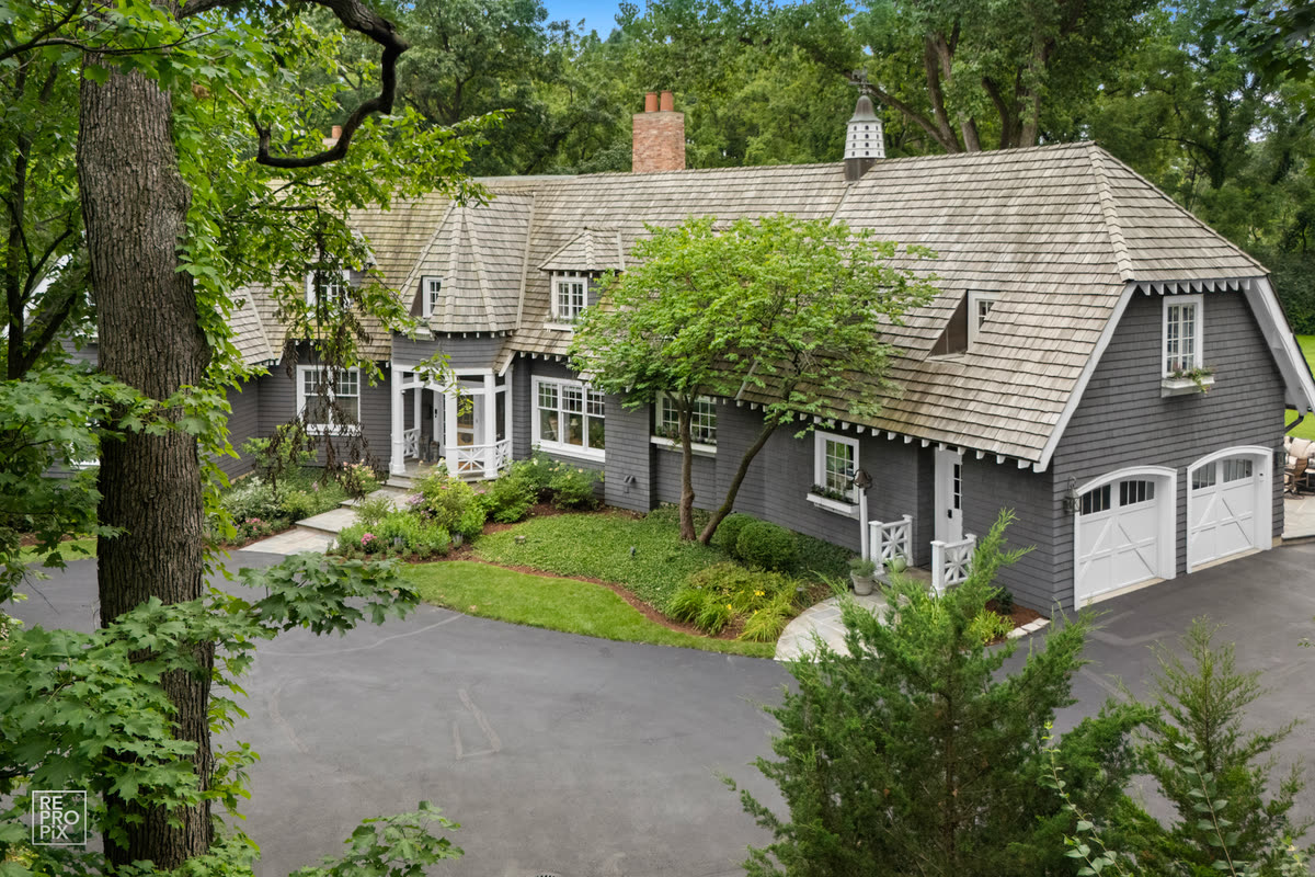 Aerial view of a charcoal-gray shingled home with white garage doors, a curved driveway and mature oaks — drone photography