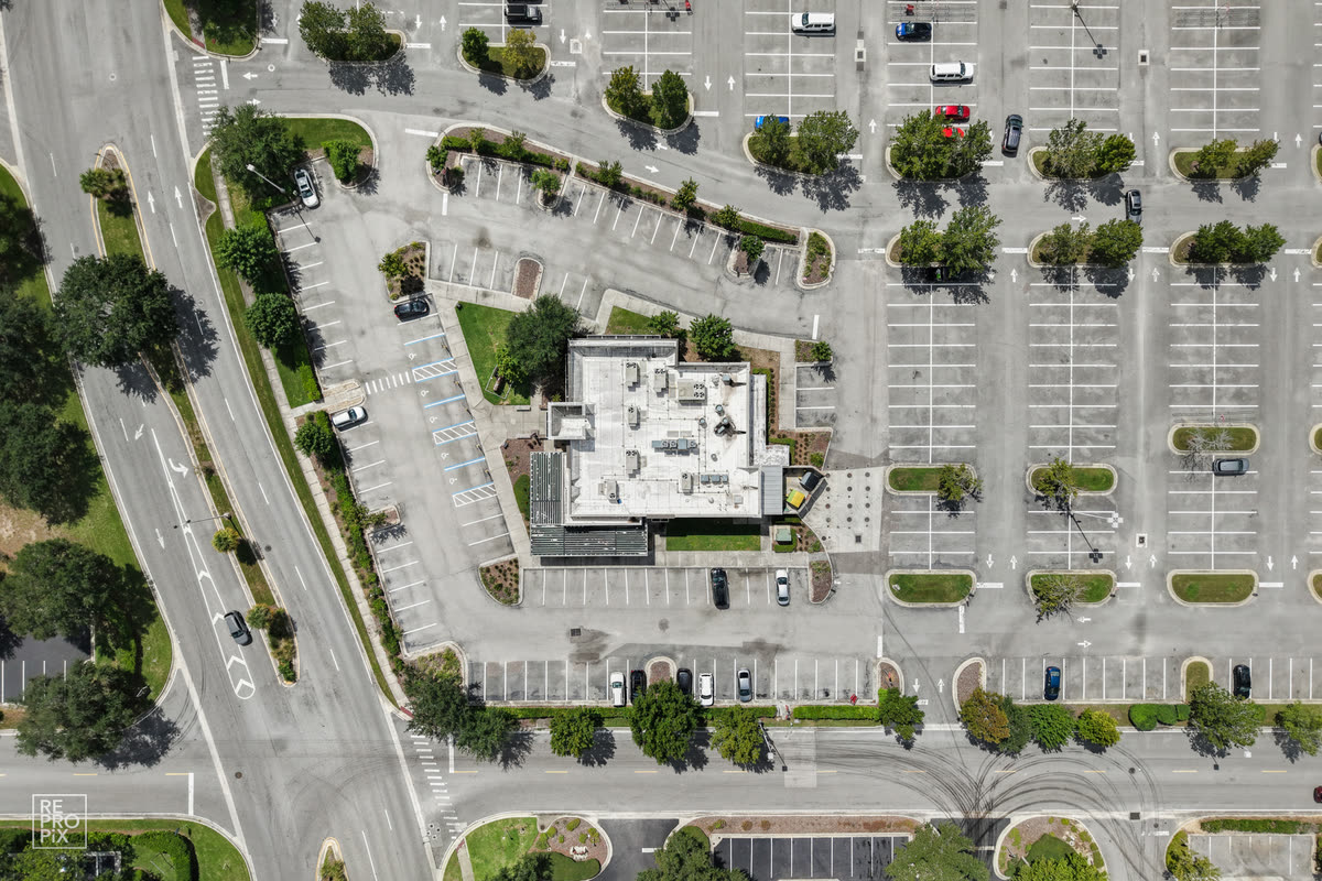 Top-down aerial view of a commercial building and its striped parking lot beside a main road — drone photography