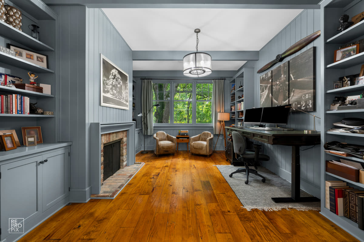 Home office with blue-gray built-in shelving, a brick fireplace and a desk beneath the window — home interior photography