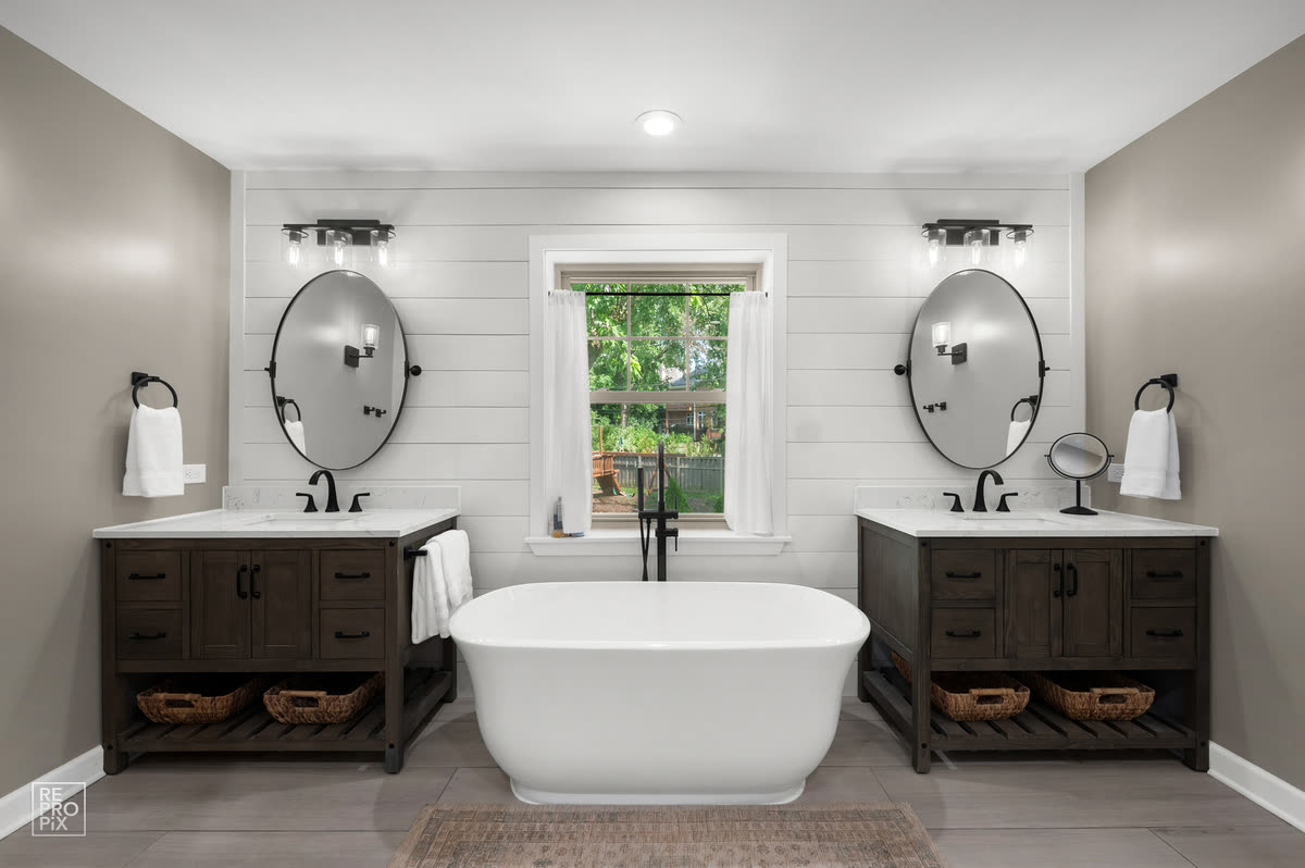 Bathroom with twin dark vanities, oval mirrors and a freestanding tub under the window — property photo
