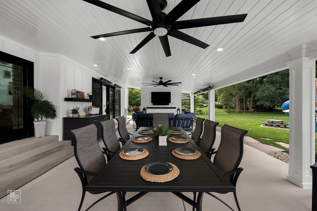 Covered porch with black ceiling fans and a long dining table set for eight, overlooking the lawn — home exterior photo