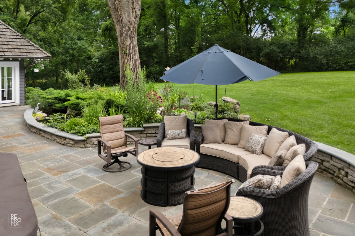 Stone patio with a curved wicker sectional, umbrella and fire table under mature trees — home exterior photo